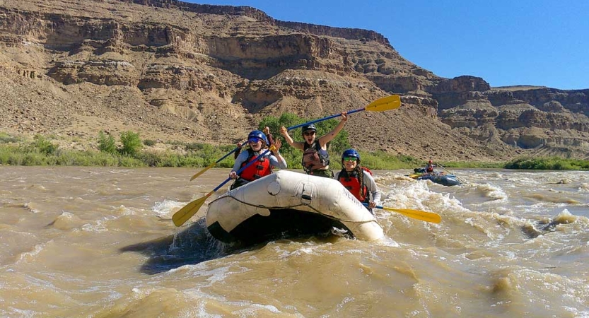 A group of people wearing life jackets and helmets navigate the choppy waters of a river. Behind them are tall canyon walls. 